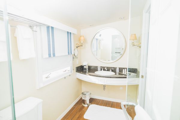 A bright, modern bathroom with a round mirror, a washbasin with a granite top, white towels, and a small trash can on a wooden floor ending the sentence.