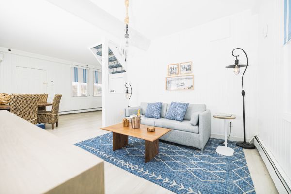 A bright living room with a gray sofa, blue rug, wooden coffee table, and wicker chairs. Light walls and blue accents seen.