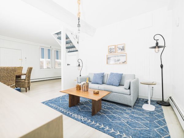 A bright living room with a gray sofa, blue rug, wooden coffee table, and wicker chairs. Light walls and blue accents seen.
