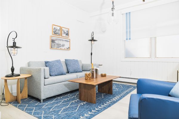 A cozy living room with a gray sofa, blue armchairs, wooden tables, and framed art, all on a blue patterned rug in a bright setting.