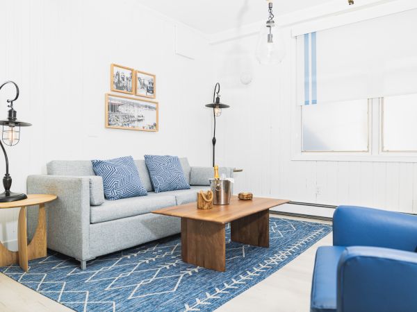 A cozy living room with a gray sofa, blue armchairs, wooden tables, and framed art, all on a blue patterned rug in a bright setting.