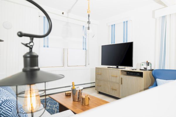 A modern living room with a TV, wooden cabinet, coffee table, and a pendulum light fixture. Light decor and striped curtains adorn the windows.