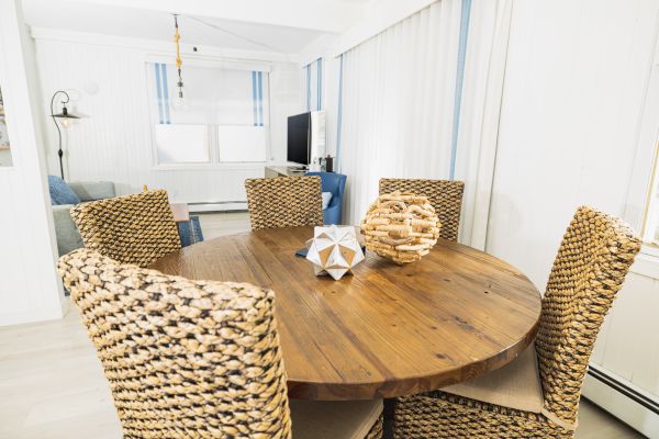 A dining area with a round wooden table, three wicker chairs, and decorative items in the center. A living area with a TV is visible in the background.