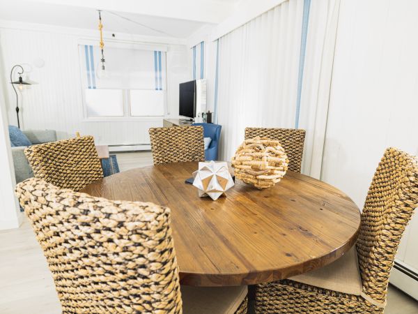 A dining area with a round wooden table, three wicker chairs, and decorative items in the center. A living area with a TV is visible in the background.