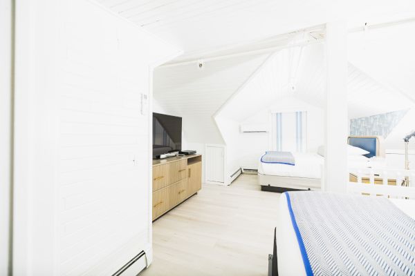 A bright, white attic bedroom with beds, a TV on a wooden stand, and sloped ceilings.