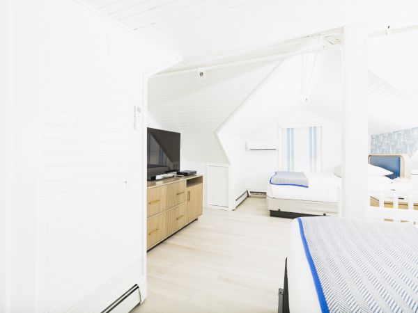 A bright, white attic bedroom with beds, a TV on a wooden stand, and sloped ceilings.