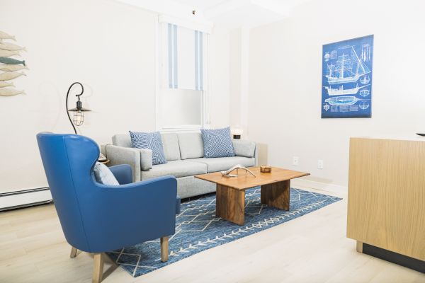 A living room with a blue armchair, grey sofa, wooden coffee table, and nautical-themed decor on a blue rug near a window and a wall painting.