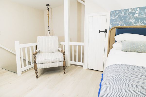 The image shows a cozy, white-themed bedroom with nautical wallpaper, a striped armchair, and a bed with blue and white bedding.