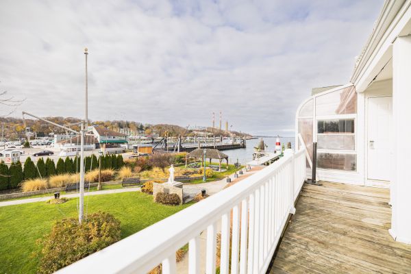 The image shows a balcony with a white railing, overlooking a green park and a harbor with boats and industrial structures under a partly cloudy sky.