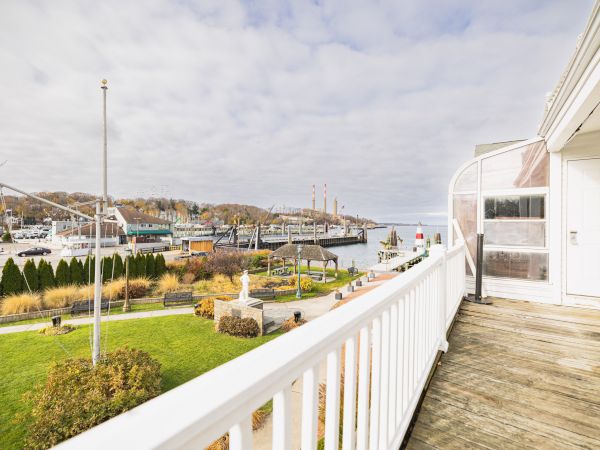The image shows a balcony with a white railing, overlooking a green park and a harbor with boats and industrial structures under a partly cloudy sky.