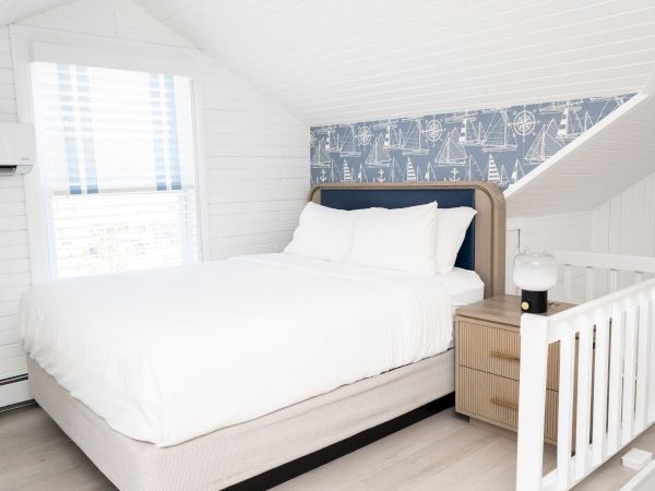 A bright white bedroom with a large bed, pillows, bedside table, and nautical sailboat wallpaper on the angled wall.