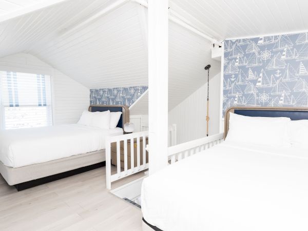Bright white bedroom loft with two beds, nautical sailboat wallpaper, a window with blinds, and a dangling light above the rail bed.