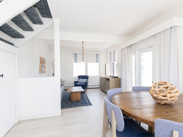 A bright living room with white walls, blue upholstered chairs, sheer curtains, a pendant light, and a wooden dining table with rope decor.