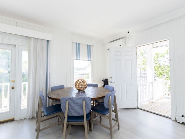 A bright dining room with a round wooden table, blue chairs, striped shades, and open doors to a deck.
