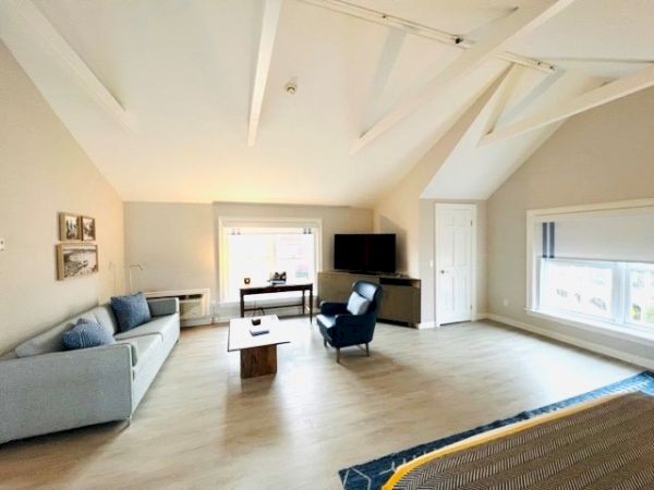 A spacious living room with a sofa, armchair, coffee table, and TV. Light-colored walls and flooring with large windows providing ample natural light.