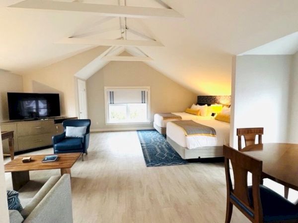 The image shows a spacious room with two beds, a TV area, a dining table, and cozy seating, all under a vaulted ceiling with exposed beams.