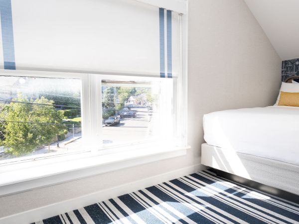 Sunlit bedroom with a bed by a window, blue-striped curtains, and a striped rug showing street traffic outside.