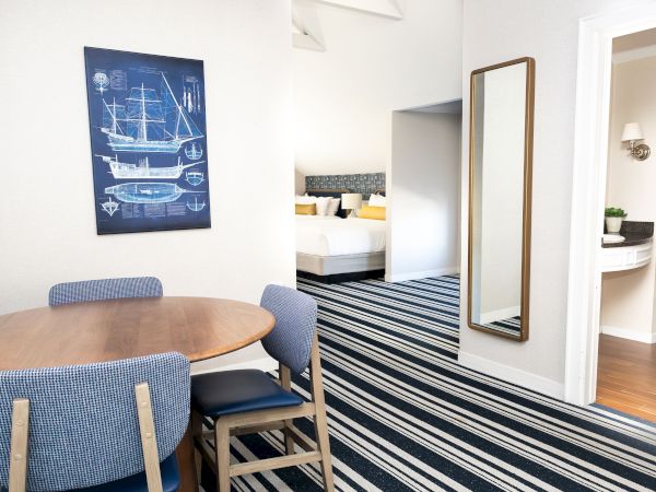 Bright hotel room: wooden round table with blue chairs, striped rug, tall gold mirror, and a blue sailboat blueprint wall art.
