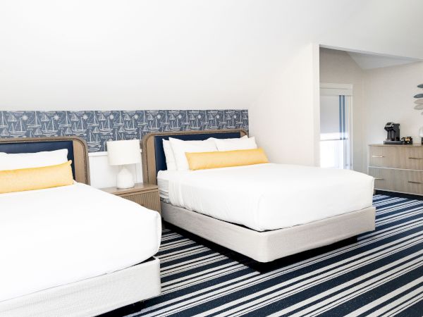 A bright nautical-themed bedroom with two made-up beds, sailboat wallpaper, striped rug, and a dresser with decor.