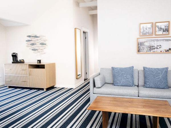 Bright living room with striped rug, gray sofa and blue pillows, framed photos, plus a cabinet with coffee maker and fish art.
