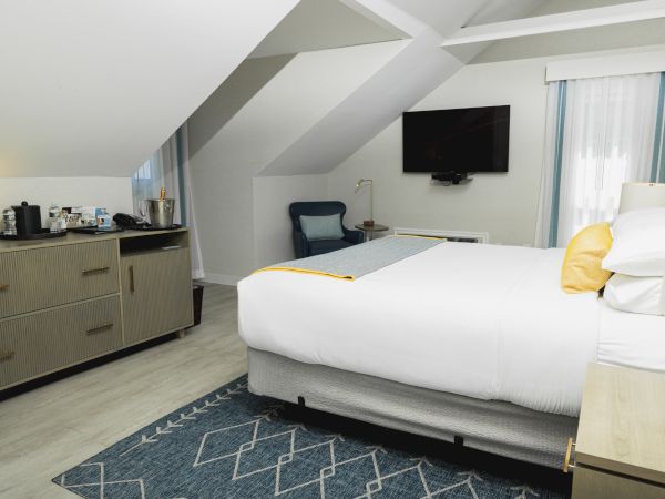 The image shows a modern hotel room with a bed, TV, dresser, and chair under an angled ceiling. There are also a coffee maker and a mini fridge.