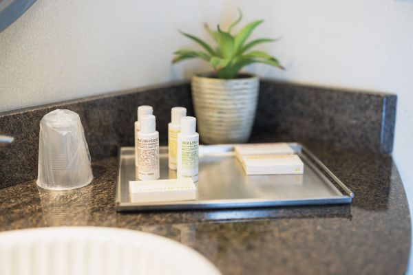 The image shows a bathroom countertop with toiletries, a wrapped cup, a small plant, and a metal tray holding bottles and boxed items.