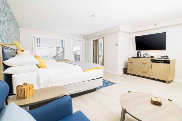 A modern hotel room with a bed, wall-mounted TV, coffee station, and seating area, featuring light-colored decor and ample natural light.