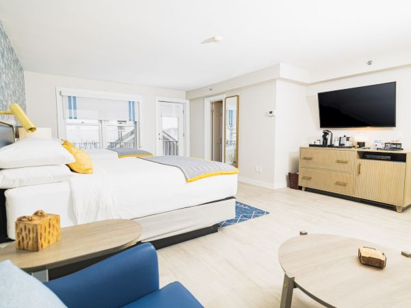 A modern hotel room with a bed, wall-mounted TV, coffee station, and seating area, featuring light-colored decor and ample natural light.