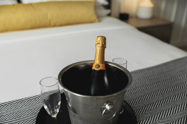 A bottle of champagne is chilling in an ice bucket with two Champagne flutes on a tray, set on a bed with a yellow pillow.