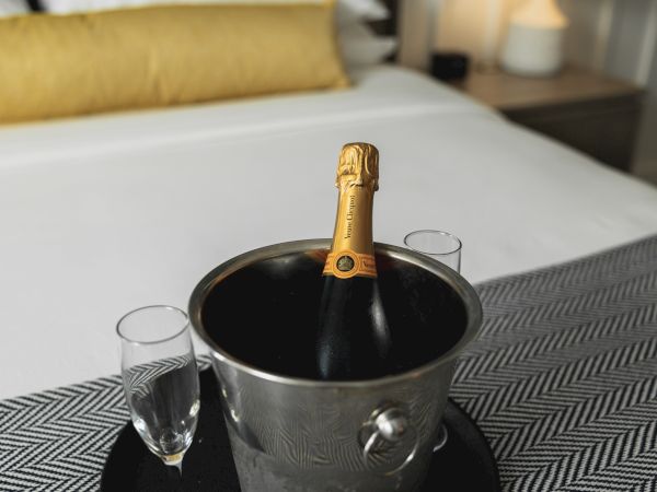 A bottle of champagne is chilling in an ice bucket with two Champagne flutes on a tray, set on a bed with a yellow pillow.
