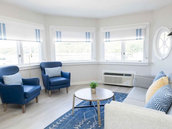 Bright living room with blue armchairs, a gray sofa, round coffee table, blue rug, and large windows with blinds.
