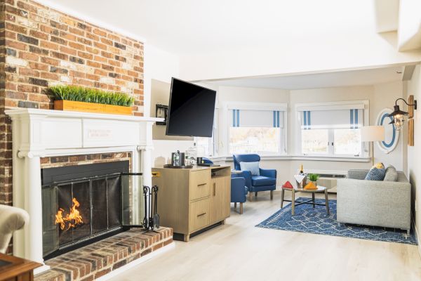 A cozy living room features a lit brick fireplace, mounted TV, blue armchairs, a gray sofa, a wooden coffee table, and large windows with striped shades.