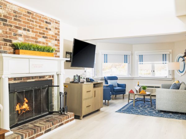 A cozy living room features a lit brick fireplace, mounted TV, blue armchairs, a gray sofa, a wooden coffee table, and large windows with striped shades.