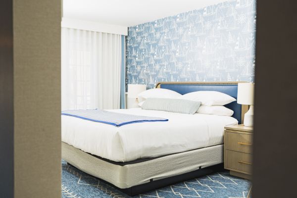 A neatly made bed with white linens in a bedroom featuring blue patterned wallpaper, bedside tables with lamps, and a window with curtains.