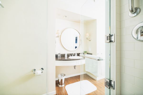 This image shows a modern bathroom with a round mirror above a sink, white walls, wooden floor, glass shower door, and towel rack.