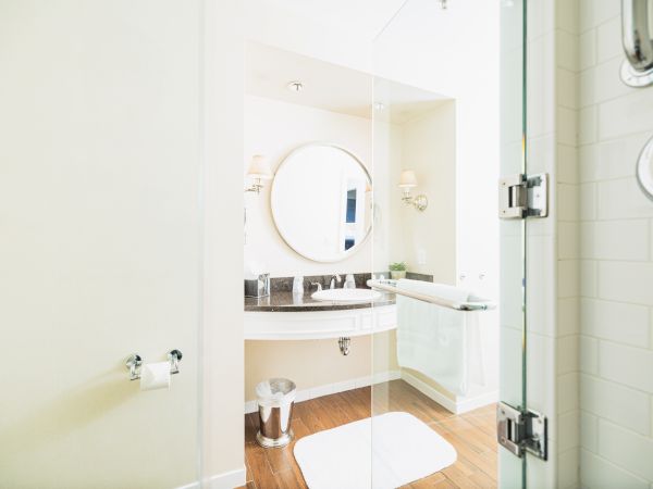 This image shows a modern bathroom with a round mirror above a sink, white walls, wooden floor, glass shower door, and towel rack.