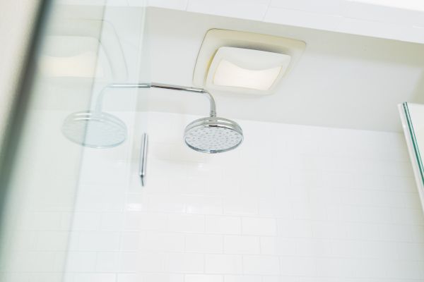 The image shows a modern shower with a glass door, a round showerhead, and a ceiling light in a bathroom with white tiled walls.