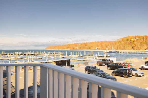 A view of a parking lot with several cars, adjacent to a marina with empty boat slips, and a scenic backdrop of a wooded shoreline and serene water.