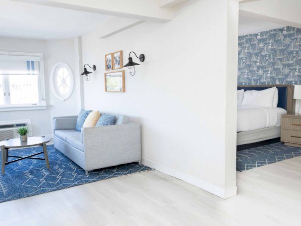 Bright coastal-style bedroom/living area with a sofa, coffee table, wall lamps, navy sailboat wallpaper, and a bed beyond.