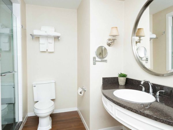 Bright modern bathroom with a toilet, glass shower, granite vanity sink, wall sconces, and a large round mirror.