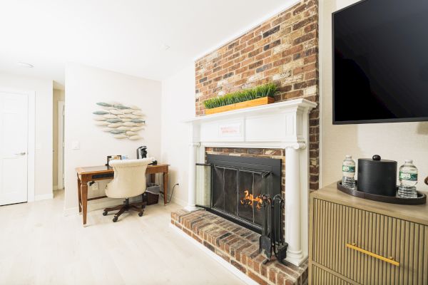 A room with a brick fireplace, a TV, a desk and chair, a wall decoration, and a cabinet with water bottles and a coffee maker.