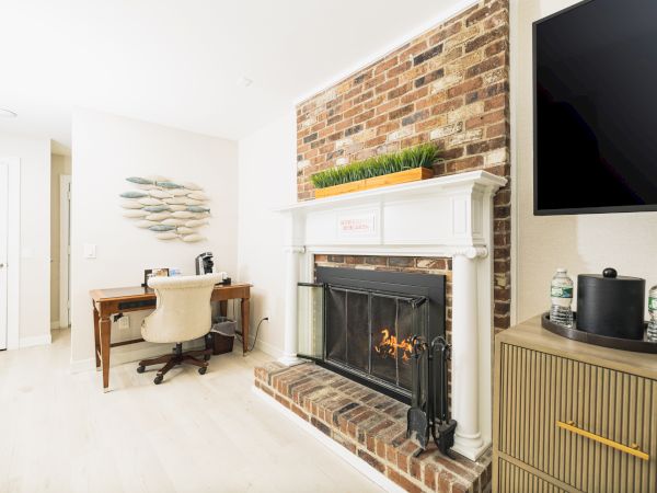A room with a brick fireplace, a TV, a desk and chair, a wall decoration, and a cabinet with water bottles and a coffee maker.