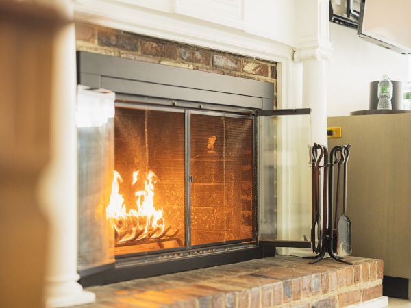 A cozy fireplace with a fire burning, metal fire tools on the side, and a modern cabinet with two bottles on top in a living room setting.