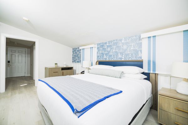 The image shows a modern bedroom with a large bed, blue and white bedding, nightstands with lamps, and a patterned blue accent wall.