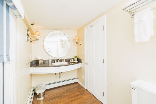 A bright bathroom with a round mirror, double sink, wall lamps, wooden floor, a wastebasket, white towels on a rack, and a white door.