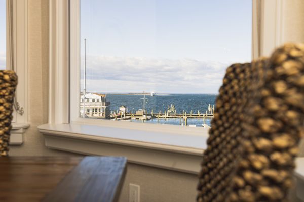 The image shows a view through a window overlooking a waterfront with boats docked at a pier, with chairs and a table in the foreground inside the room.