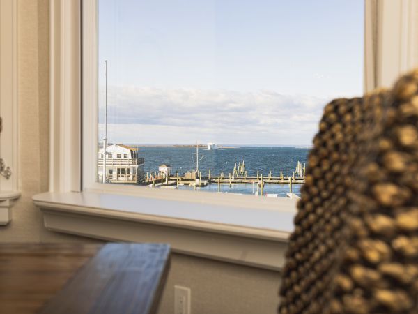 The image shows a view through a window overlooking a waterfront with boats docked at a pier, with chairs and a table in the foreground inside the room.