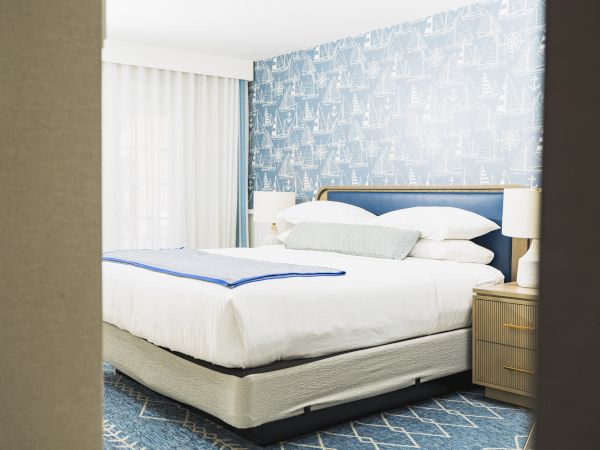The image shows a cozy bedroom with a large neatly made bed, blue-themed wallpaper, bedside tables, and curtains partially covering the window.
