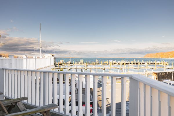 A seaside view from a white deck railing, overlooking a marina with boats and calm water, under a blue sky with scattered clouds, ending the sentence.