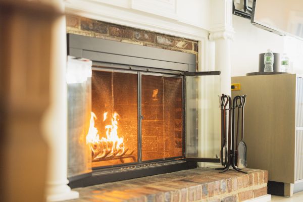 A cozy fireplace with a lit fire, surrounded by a brick hearth. Fireplace tools and a cabinet with items on top are on the right side.